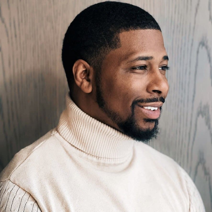 Portrait of Kareem Talbert in a cream turtleneck, smiling, against a pale wood wall